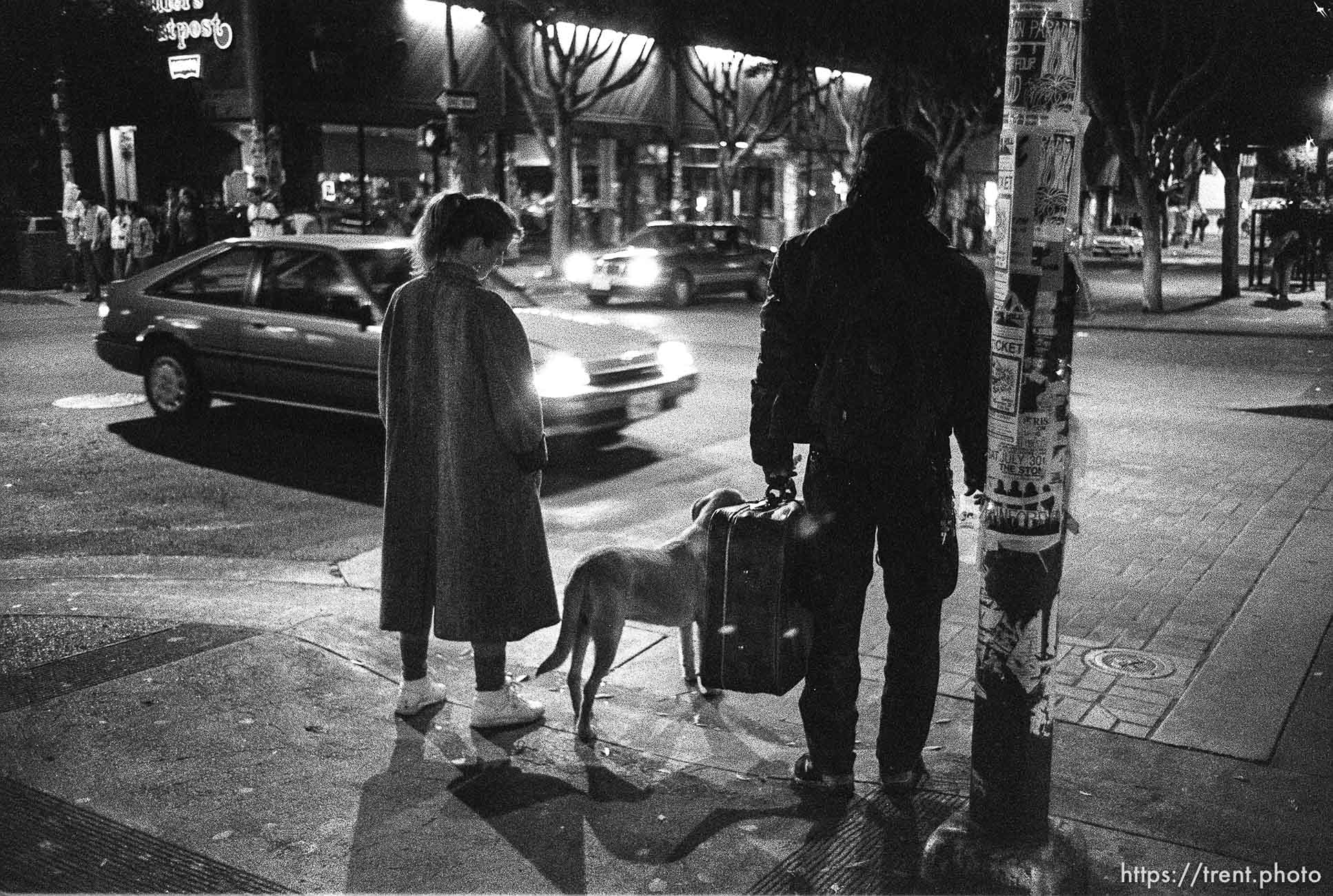 Man with suitcase and woman with dog on Telegraph Avenue at night.