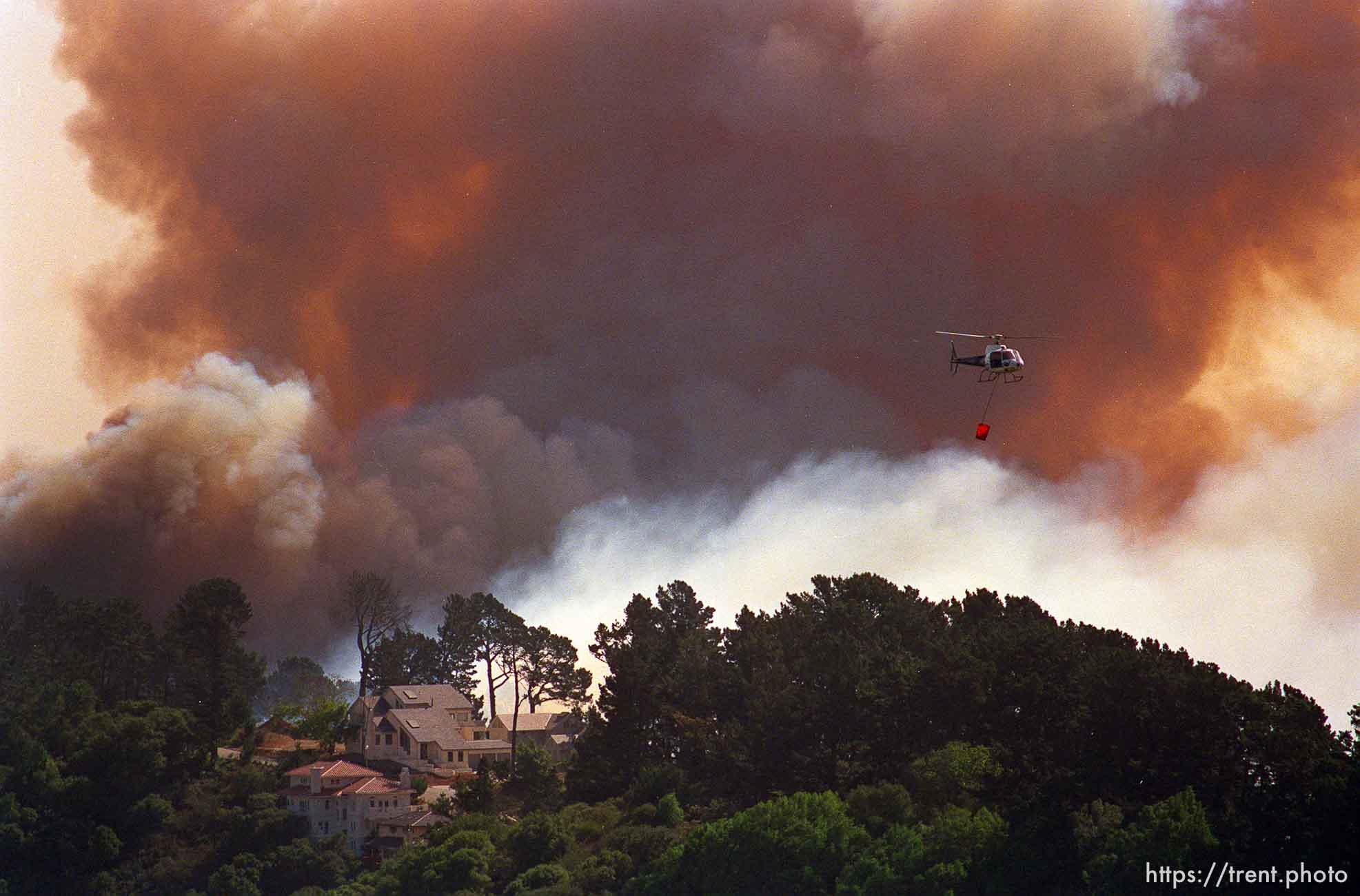 The Oakland Hills Fire threatens homes on Grizzly Peak while a helicopter makes water runs. The homes survived.