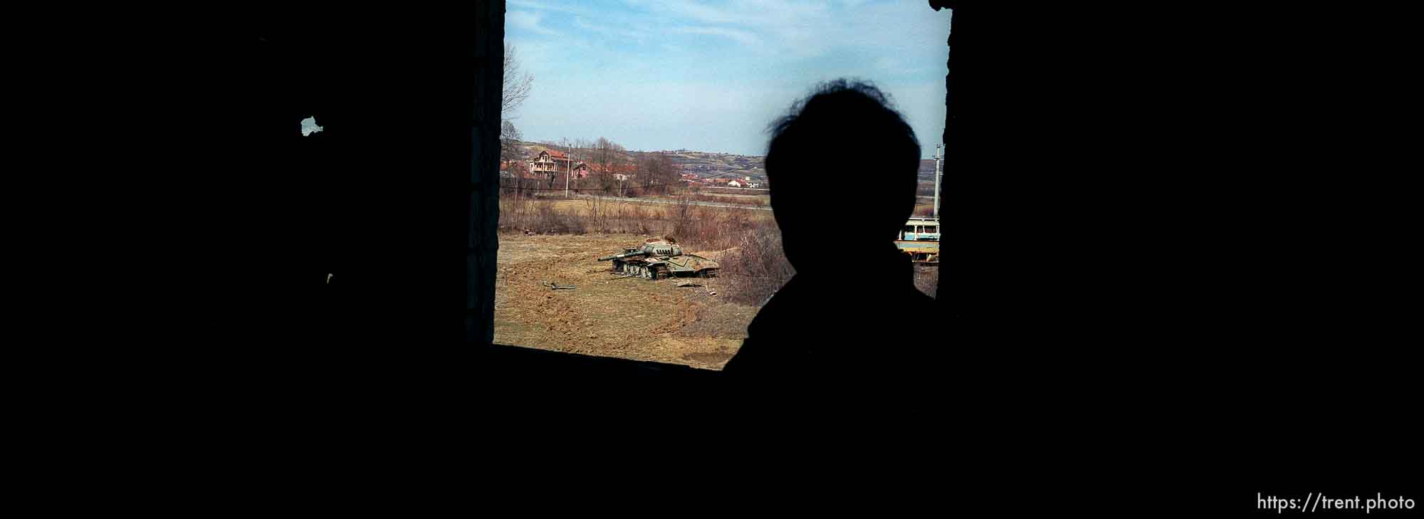 Naim Begiri looking out of a window of his uncle's home, where he hid from Serbs during the war. Outside is a destroyed Yugoslav tank.