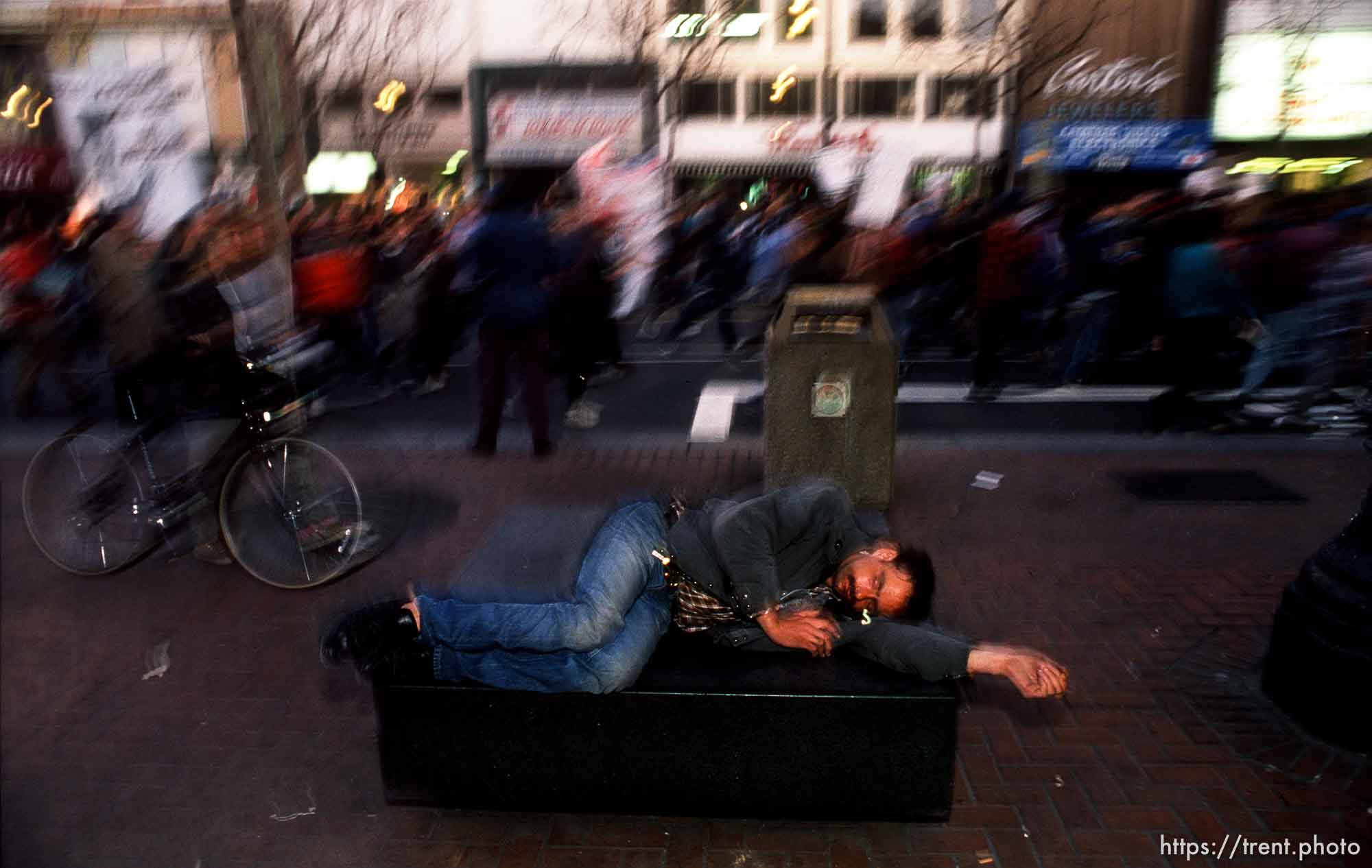 Homeless man on a bench as parade of protesters passes him on Market Street during Anti-war Gulf War protests.